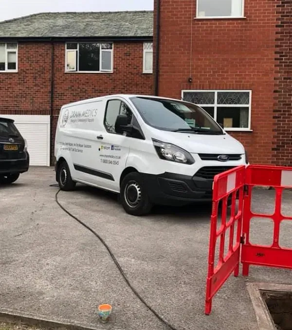 A white drainage service van parked in front of a brick house, with a hose leading to an open manhole surrounded by red safety barriers, indicating drain unblocking or repair work.