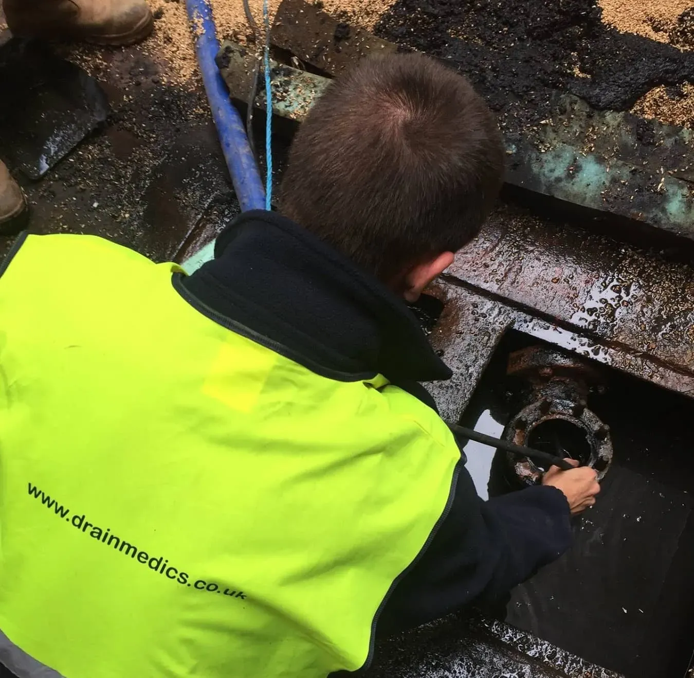 Close-up of a drain unblocking specialist in a neon yellow safety vest using a tool to clear a dirty drain opening.