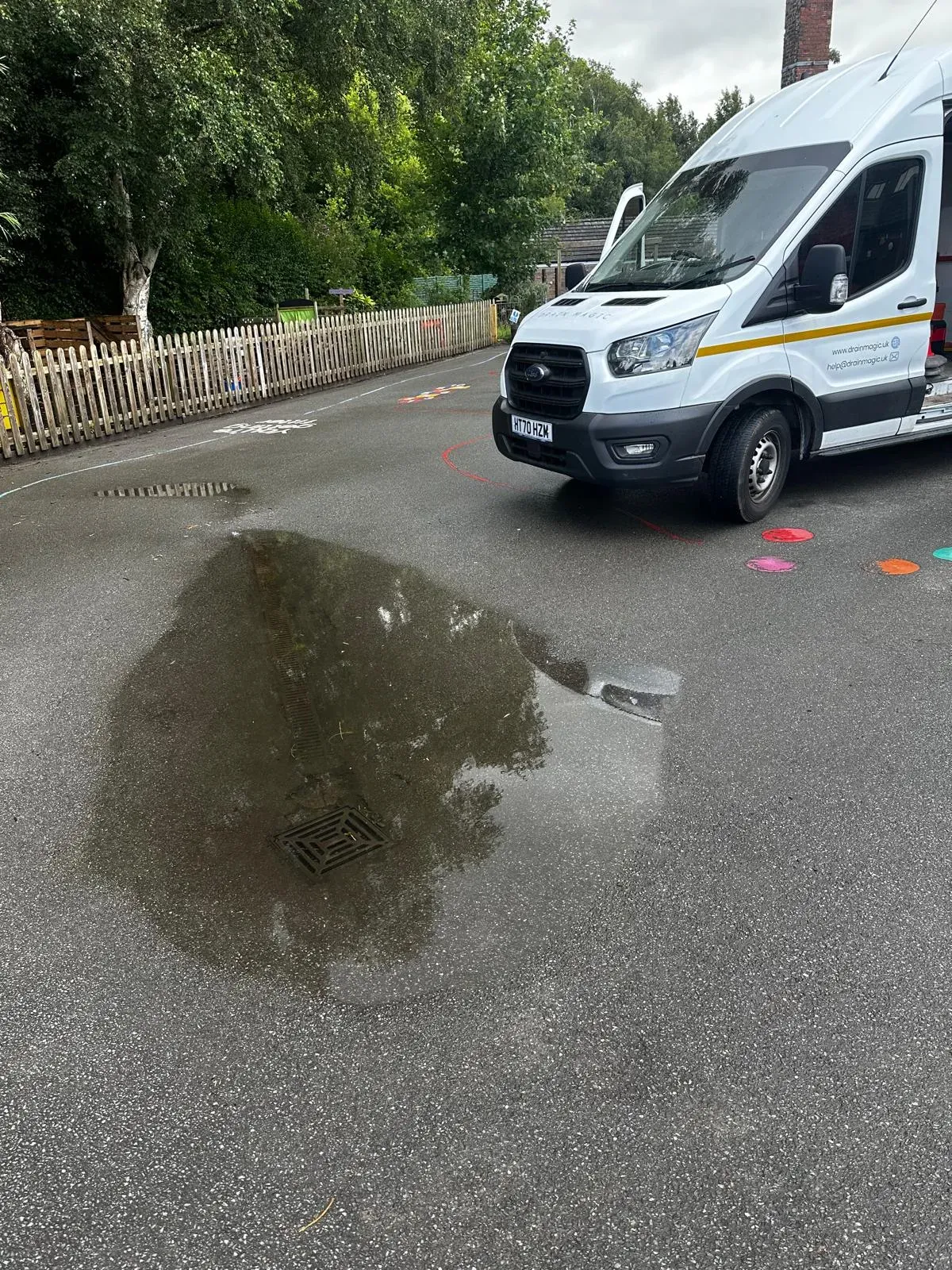 An outdoor blocked drain causing a large puddle on tarmac, with a white service van ready to clear the blockage, suggesting an emergency call-out.