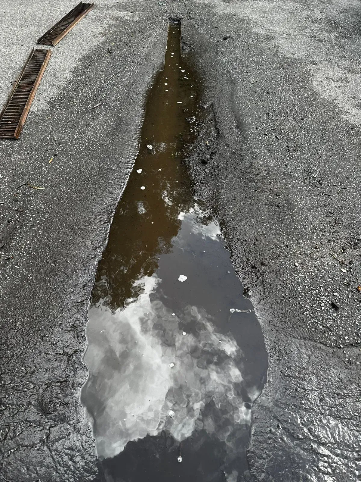 Overflowing external drain or gully with dirty water and two displaced metal grates, reflecting clouds.