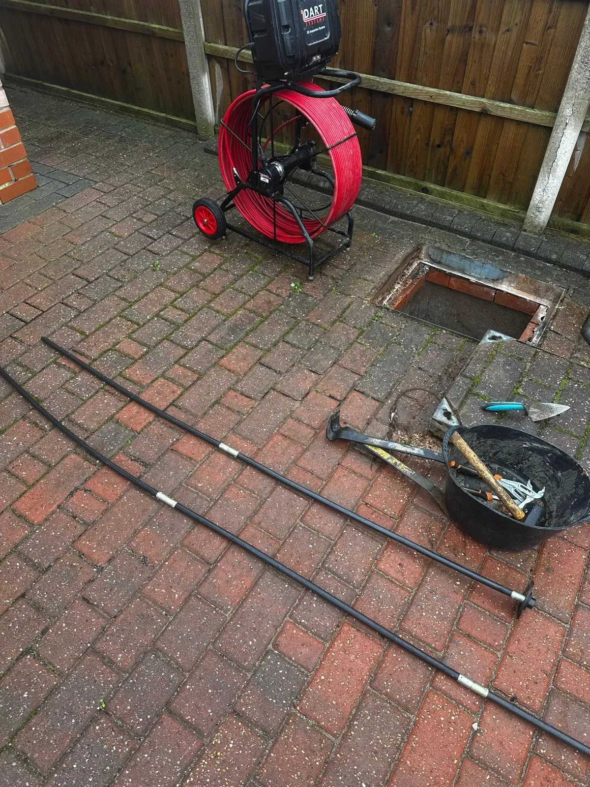 CCTV drain camera reel, drain rods, and tools next to an open manhole on a paved area, signifying a drainage survey or unblocking.