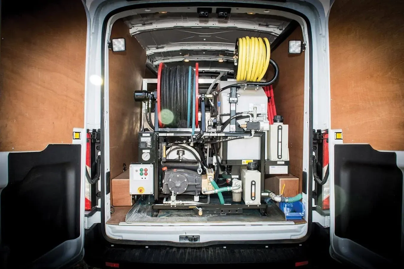 Rear view of a fully equipped white drain unblocking van with high-pressure water jetting machinery, black and yellow hoses on reels, and control panels inside.
