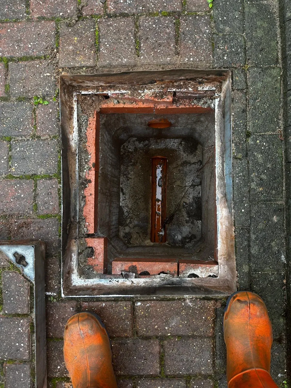 A picture looking down into an open drain manhole, showing standing water from a blockage, with a worker's boots in the foreground.