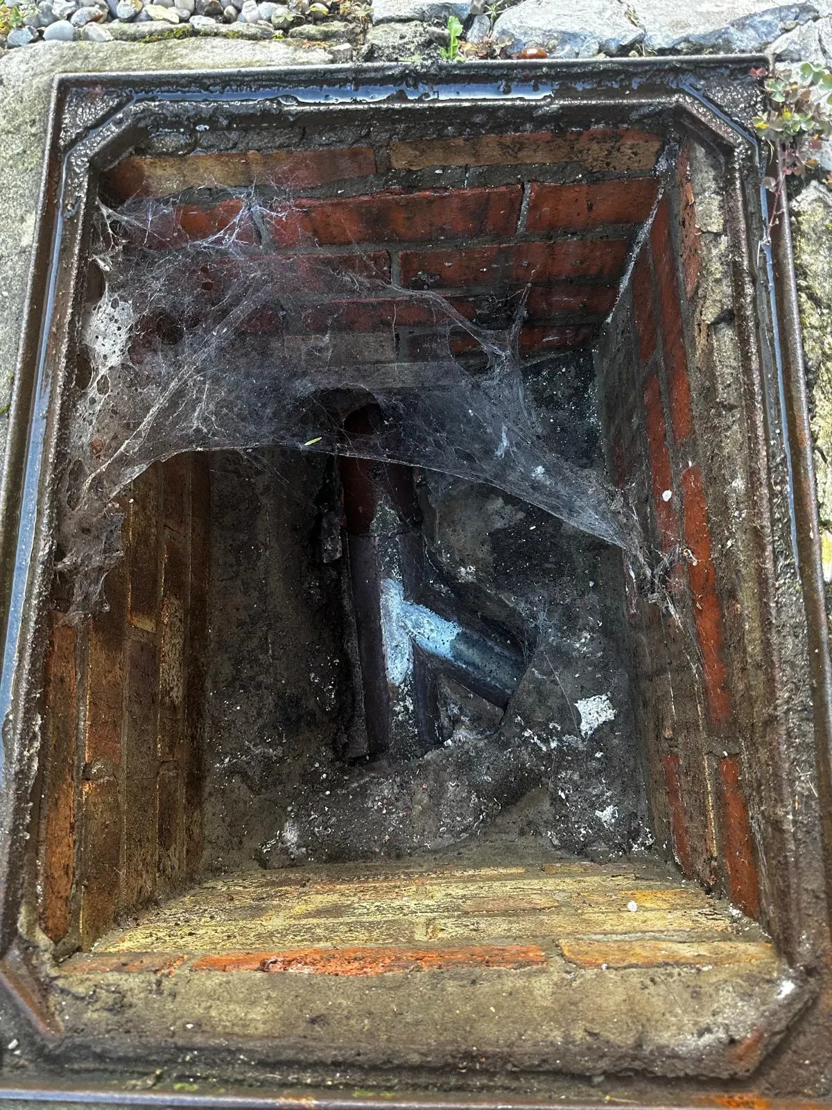 A residential drain inspection chamber or manhole, dirty and with cobwebs, used for accessing blocked or damaged drainage systems.