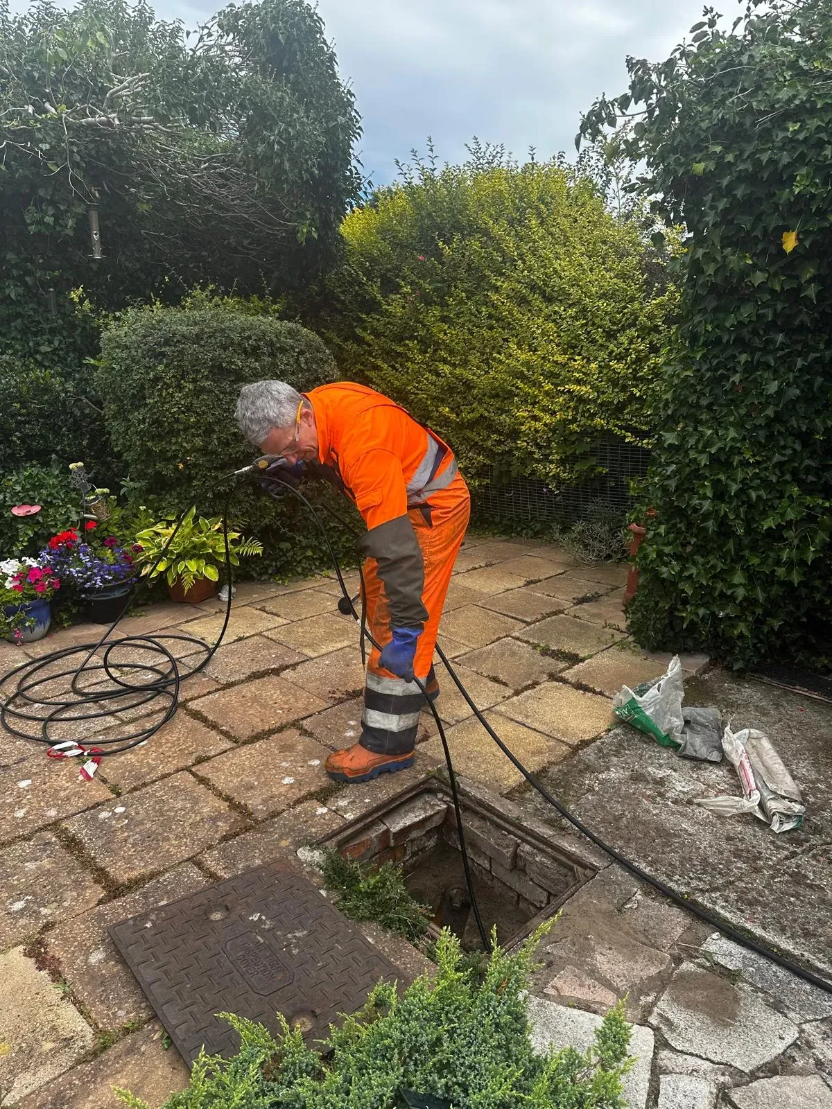 A drain unblocking technician examining an outdoor drain in a garden, with equipment for clearing blockages or conducting a CCTV survey.
