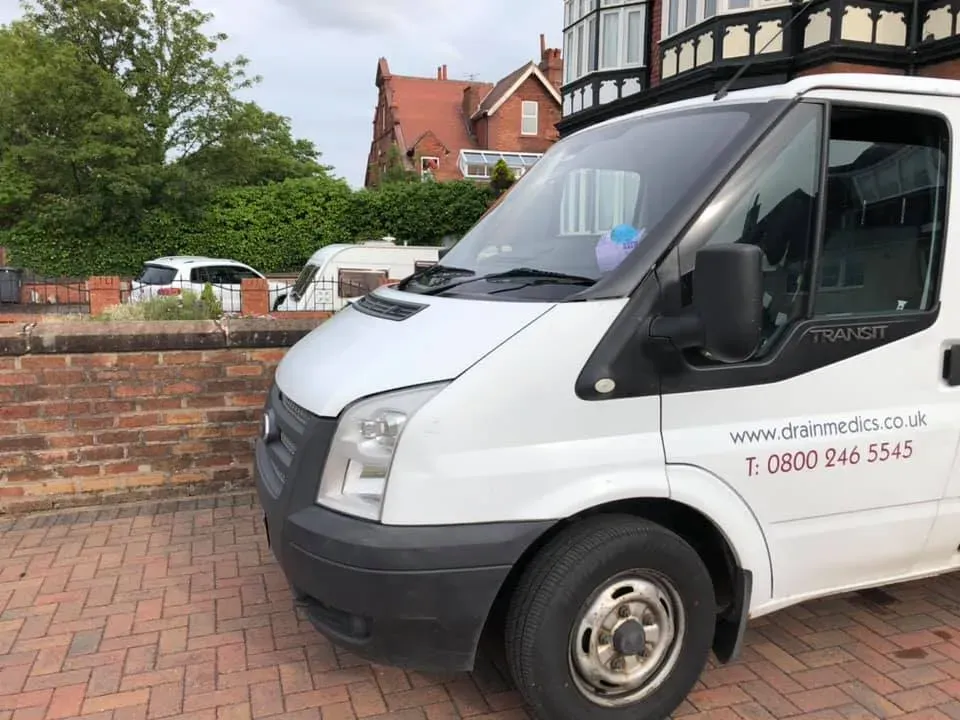 A white drainage service van parked on a paved driveway with a brick wall and residential building in the background.