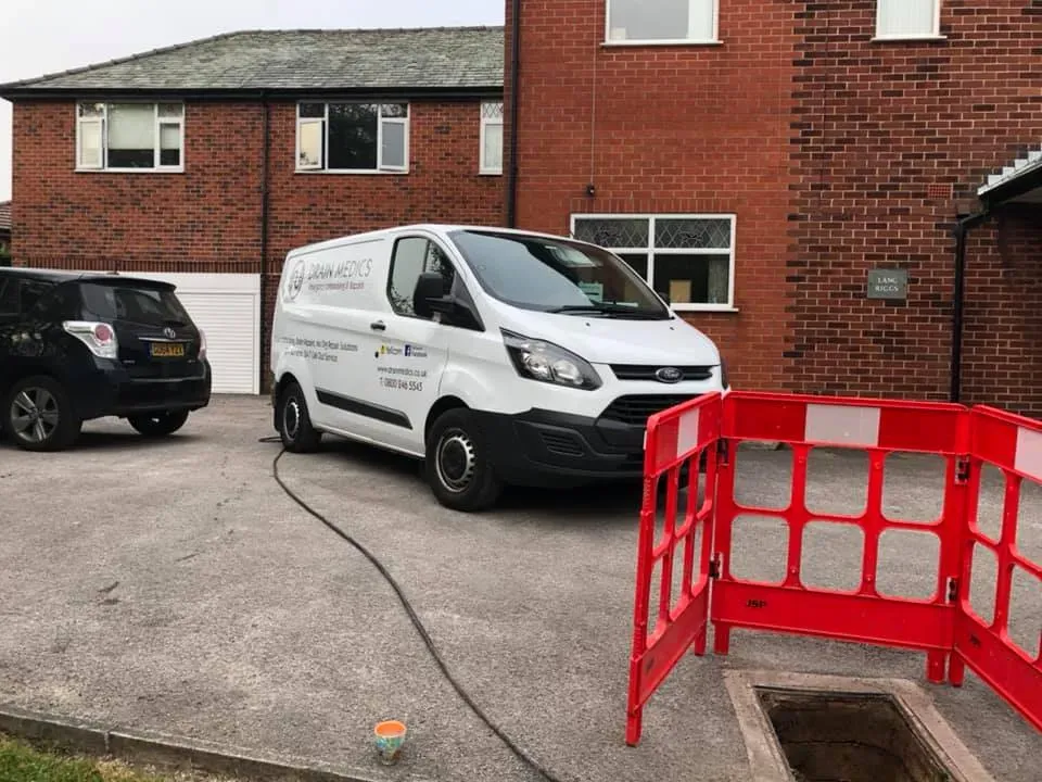 A white drainage service van, an open manhole with red safety barriers, and a residential brick house in the background, indicating professional drain services.