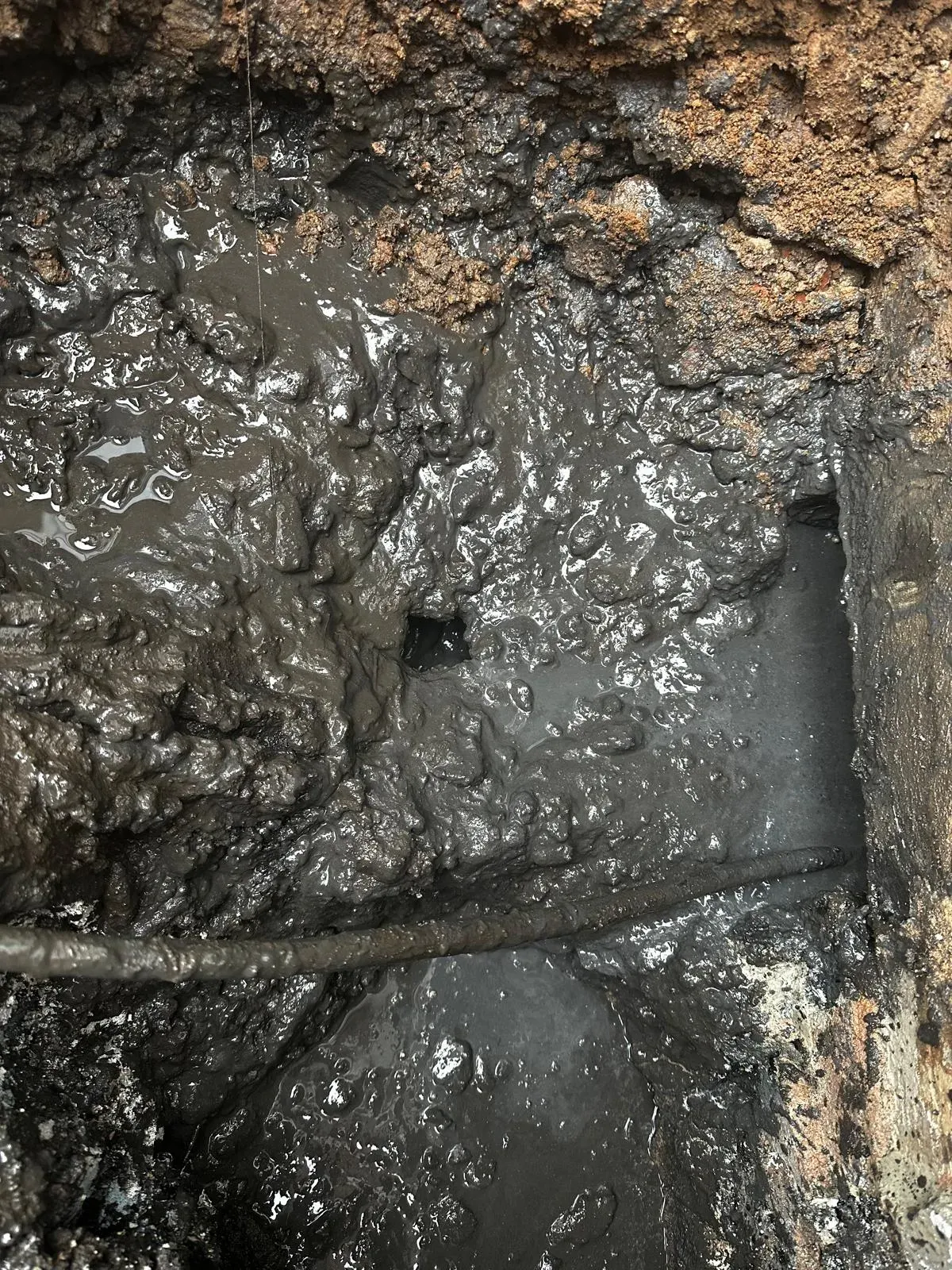 A close-up view of a severely blocked drain, overflowing with dark, greasy sludge and foul water, with a drain rod being used to clear the blockage.