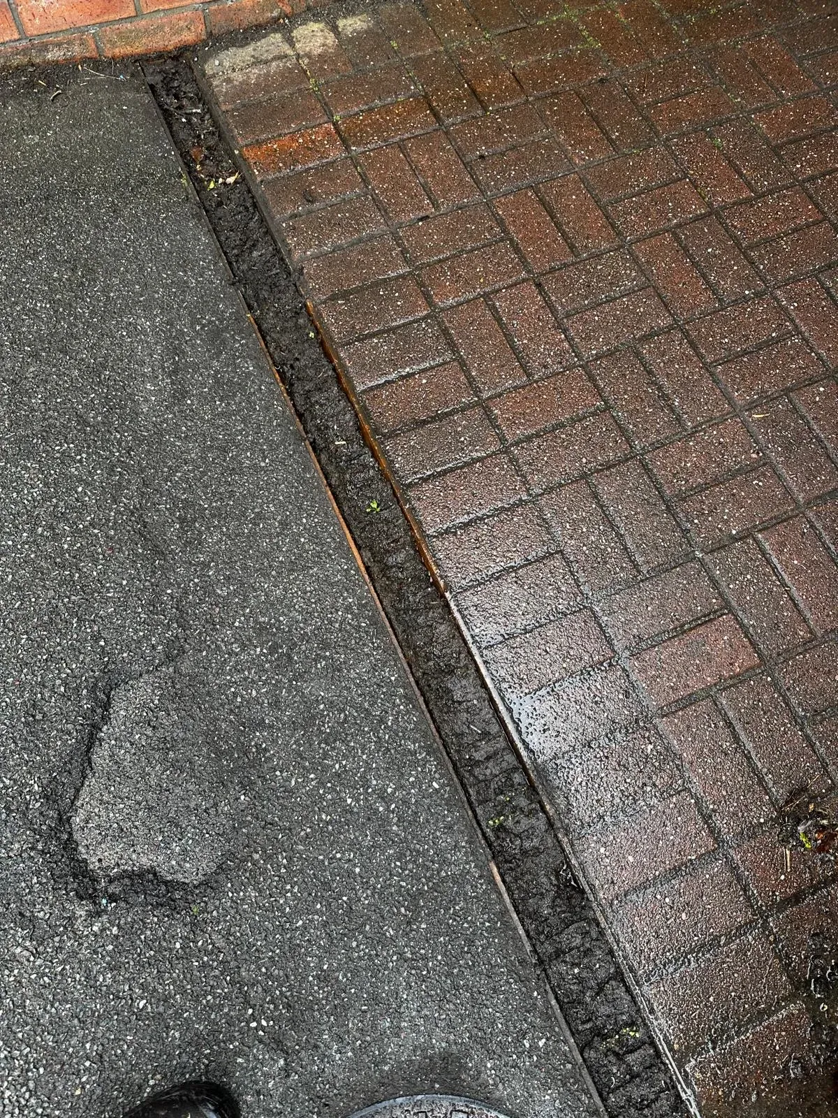 Blocked outdoor drainage gully filled with debris between a tarmac path and brick paving, highlighting the need for professional drain unblocking services in Skipton.
