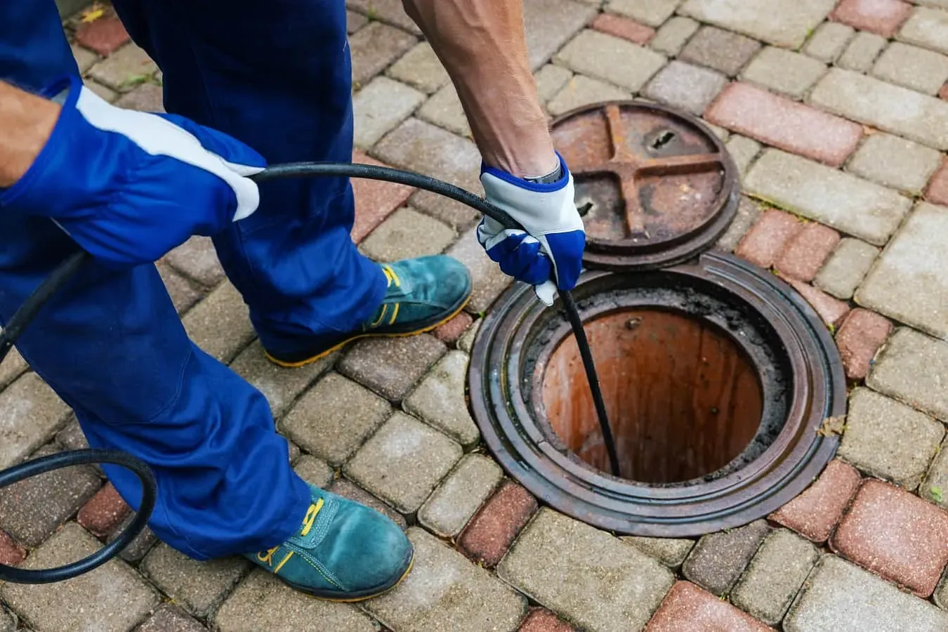 A professional drain unblocking specialist inserting a tool into an open manhole on a paved surface.